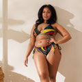 Eine Frau mit dunklen, gelockten Haaren präsentiert einen Triangle-Bikini mit einem farbenfrohen, abstrakten Allover-Print. Das Design kombiniert kräftiges Gelb, Rot, Türkis und Orange auf schwarzem Grund in einem dynamischen Wirbel-Muster. Das Oberteil und die tief sitzende Hose sind mit Bändern zum Schnüren versehen. Die Pose ist selbstbewusst vor einem neutralen, hellen Hintergrund mit leichtem Schattenwurf.